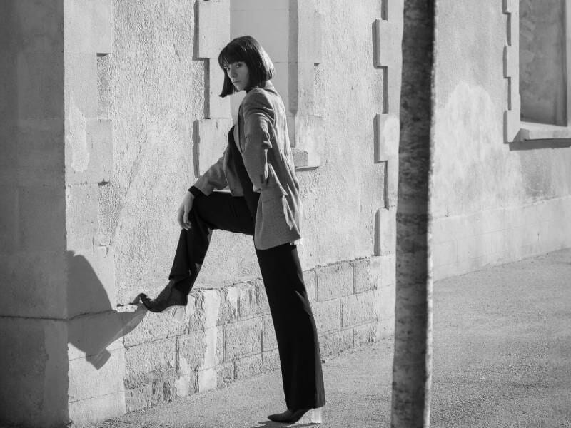 Femme élégante posant contre un mur en pierre, une jambe levée, vêtue d'un pantalon noir et d'un blazer clair.