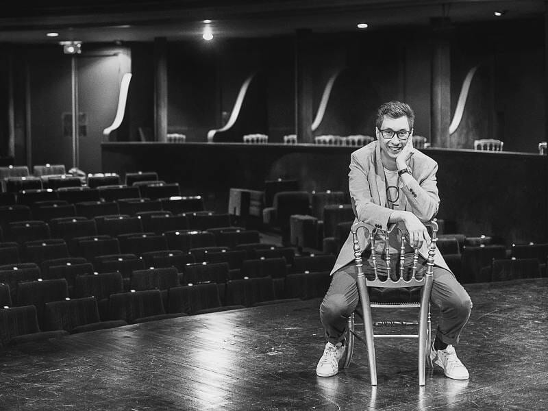 Homme assis sur une chaise sur scène dans un théâtre vide, souriant et portant des lunettes et des baskets blanches.