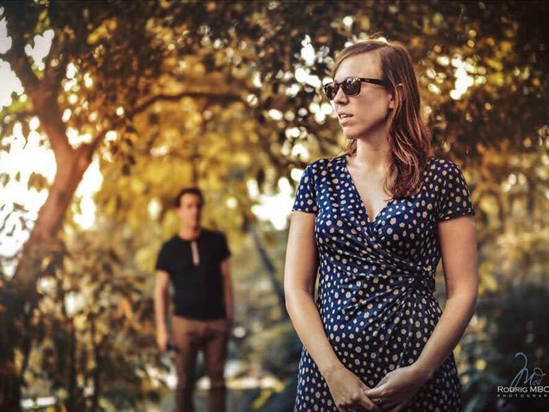 Femme en robe bleue à pois blancs et lunettes de soleil, homme flou en arrière-plan lors d'un shooting photo lifestyle au Mans.