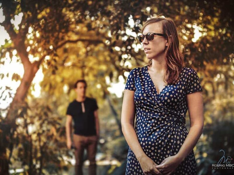 Femme en robe bleue à pois blancs et lunettes de soleil, homme flou en arrière-plan lors d'un shooting photo lifestyle au Mans.