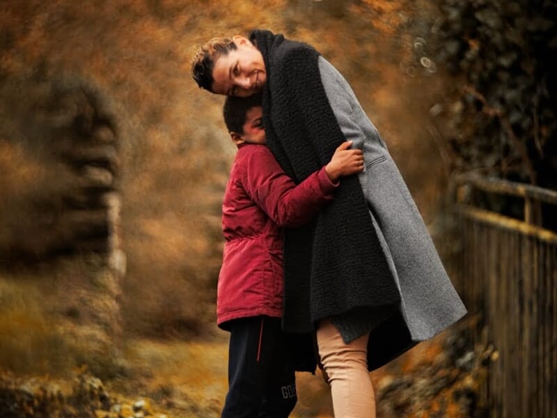 Une femme en manteau gris serre dans ses bras un enfant en veste rouge dans un décor automnal lors d'un shooting photo famille au Mans.