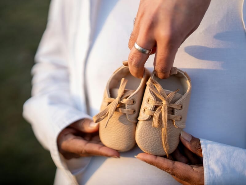 Une personne tenant une paire de petites chaussures beige devant un ventre de femme enceinte lors d'un shooting photo bébé au Mans.