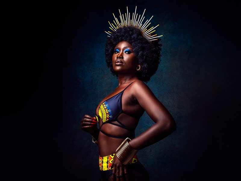 Portrait d'une femme noire avec une coiffure afro et un ornement doré, portant un vêtement coloré et un maquillage bleu.