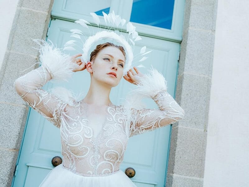 Femme blanche en robe blanche ornée de plumes, posant devant une porte bleue avec un halo de plumes autour de la tête.