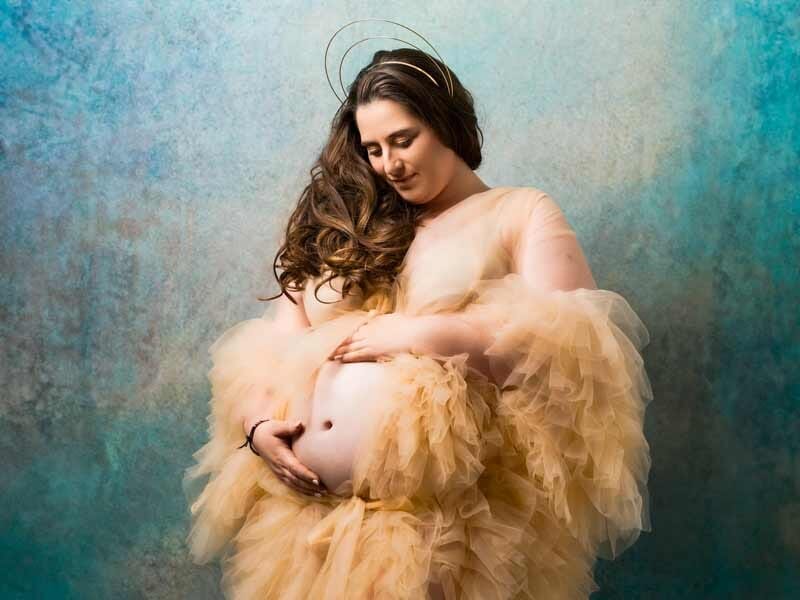 Femme enceinte en robe beige vaporeuse tenant son ventre, fond bleu artistique, ambiance douce et sereine.