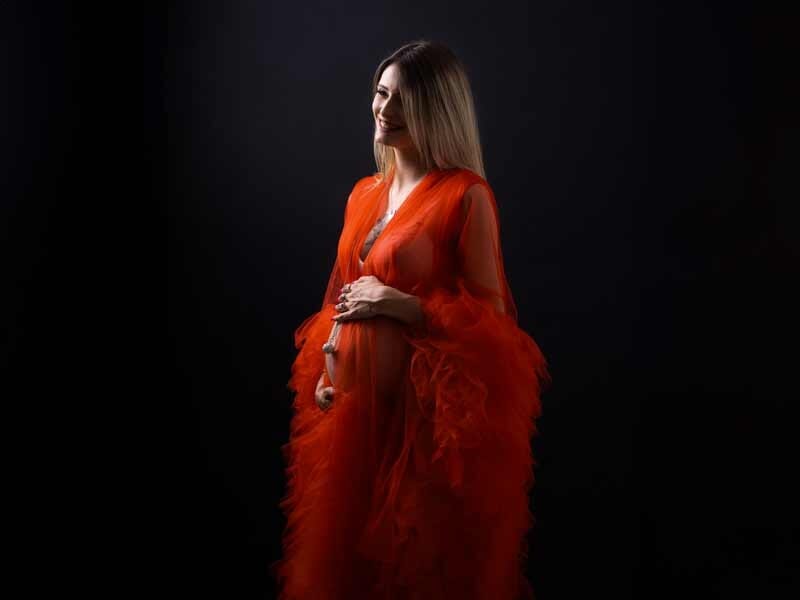 Femme enceinte en robe rouge transparente avec volants, posant de profil sur fond noir, lumière douce.