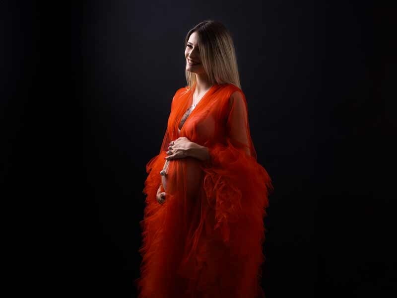 Femme enceinte en robe rouge transparente avec volants, posant de profil sur fond noir, lumière douce.
