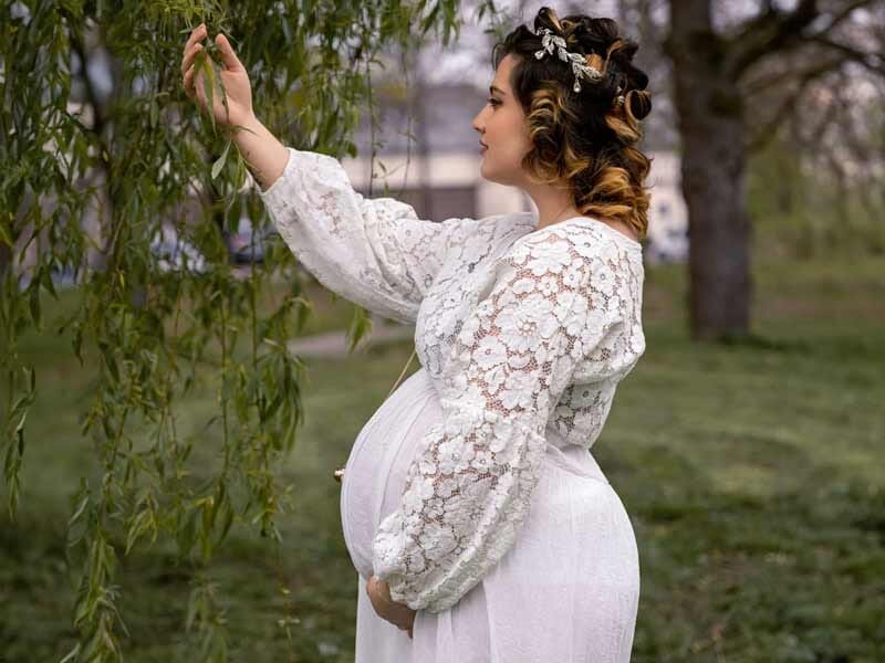 Femme enceinte en robe blanche en dentelle, debout dans un jardin, touchant une branche d'arbre.