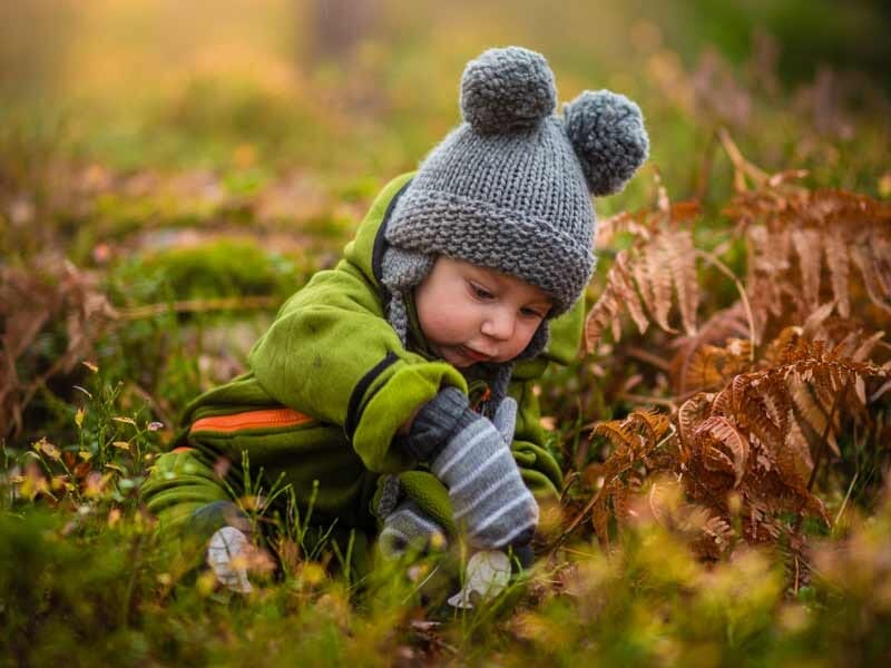 Bébé en tenue verte et bonnet gris avec pompons, jouant dans l'herbe et les feuilles en automne.