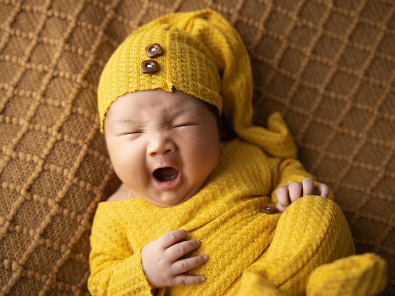 Bébé bâillant, habillé en jaune avec bonnet et combinaison, allongé sur un tissu marron texturé.
