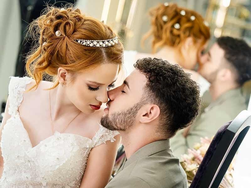 Jeune couple en tenue de mariage, s'embrassant tendrement devant un miroir dans une pièce lumineuse.