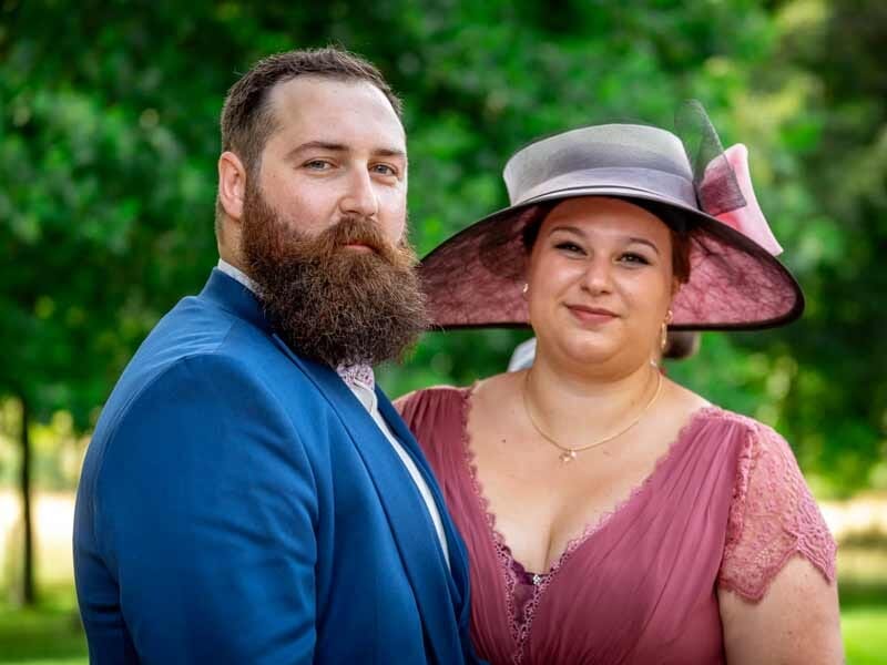 Un homme barbu en costume bleu et une femme en robe rose avec un grand chapeau posent dans un cadre naturel.