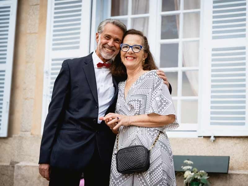 Couple souriant habillé élégamment, posant devant une maison avec des volets blancs.