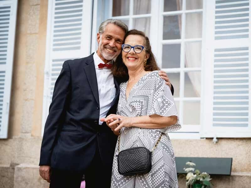 Couple souriant habillé élégamment, posant devant une maison avec des volets blancs.