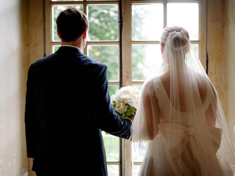 Marié et mariée vus de dos, tenant un bouquet, regardant par une fenêtre ancienne.