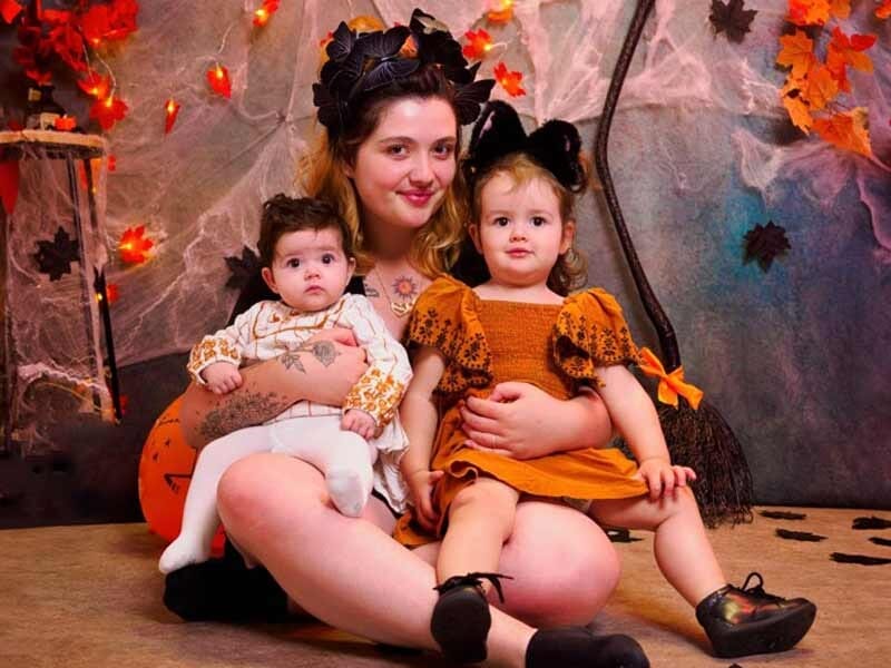Jeune femme tenant deux enfants déguisés pour Halloween, décor automnal avec citrouilles et toiles d'araignée.