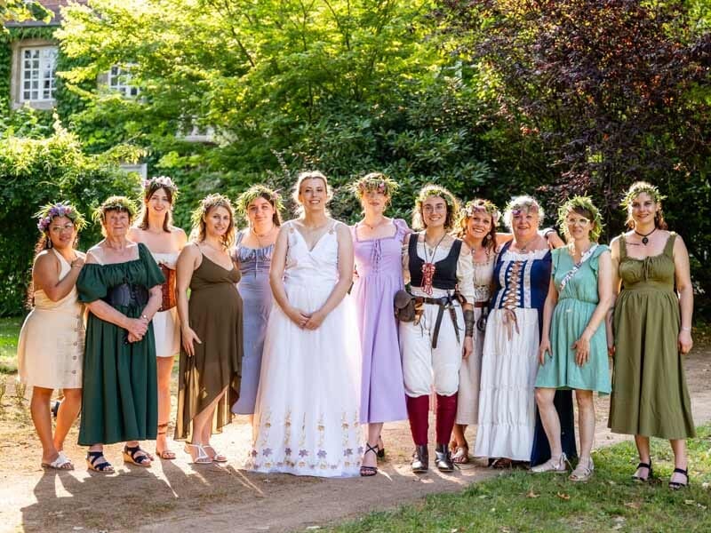 Groupe de femmes en robes colorées et couronnes de fleurs, posant en extérieur sous un arbre verdoyant.