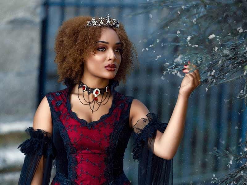 Femme en robe rouge et noire avec couronne, posant près d'un arbre aux feuilles argentées en extérieur.