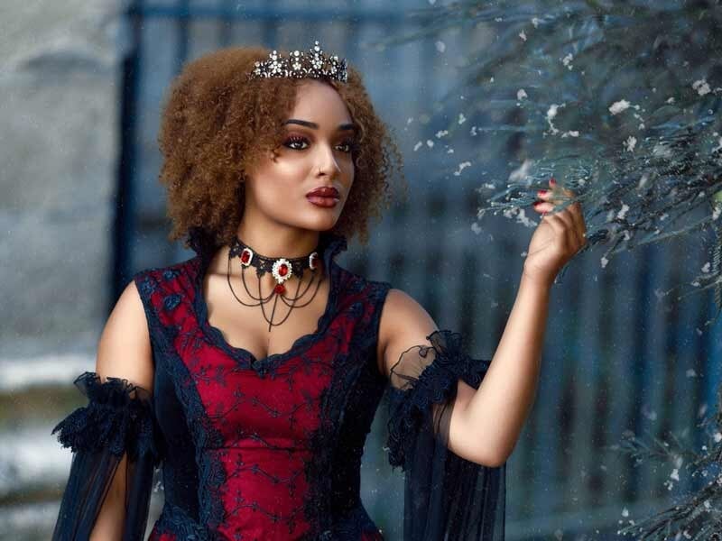 Femme en robe rouge et noire avec couronne, posant près d'un arbre aux feuilles argentées en extérieur.