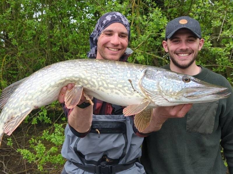 Deux hommes souriants montrent un grand brochet fraîchement pêché dans la rivière Aube, devant un fond de feuillage vert.