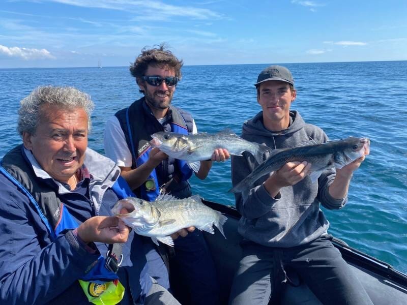 Trois pêcheurs souriants sur un bateau en mer tenant chacun un poisson fraîchement pêché sous un ciel clair.