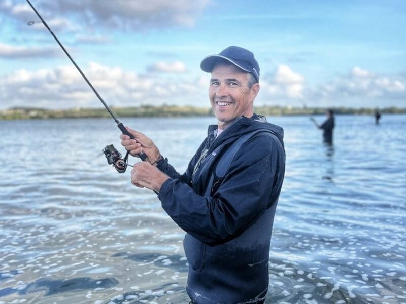 Homme souriant en tenue de pêche avec casquette, tenant une canne à pêche lors d'un stage de pêche à Saint-Pol-de-Léon.