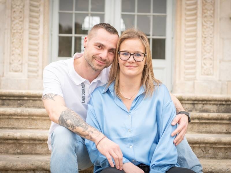 Un couple assis sur des marches devant un bâtiment ancien, l'homme enlace la femme avec un sourire doux.