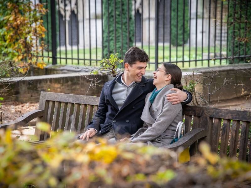 Un couple assis sur un banc en bois dans un parc, échangeant un regard complice et souriant, lors d'un shooting photo près de Rouen.