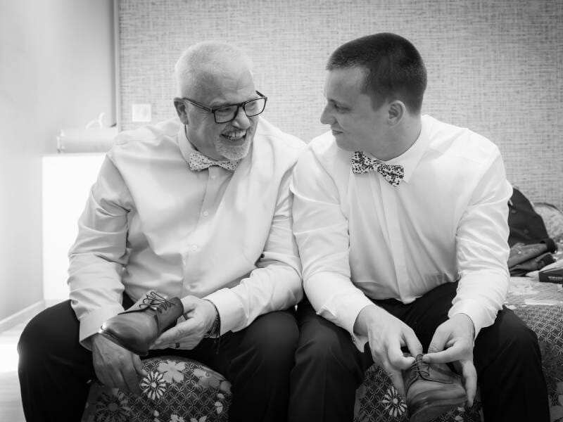 Deux hommes souriants en chemises blanches et nœuds papillon, assis et tenant des chaussures dans une pièce.