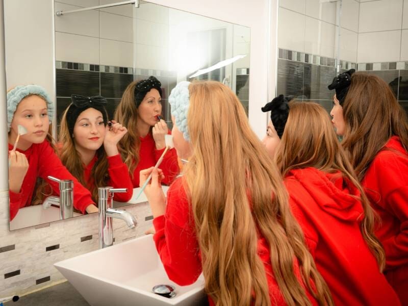 Deux jeunes filles et une femme en sweat rouge se maquillent devant un miroir dans une salle de bain moderne, lors d'un shooting photo près de Rouen.