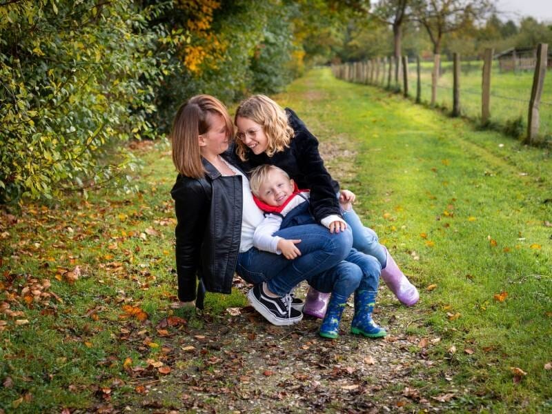 Une femme et deux enfants souriants assis sur un chemin en pleine nature avec des arbres et une clôture.