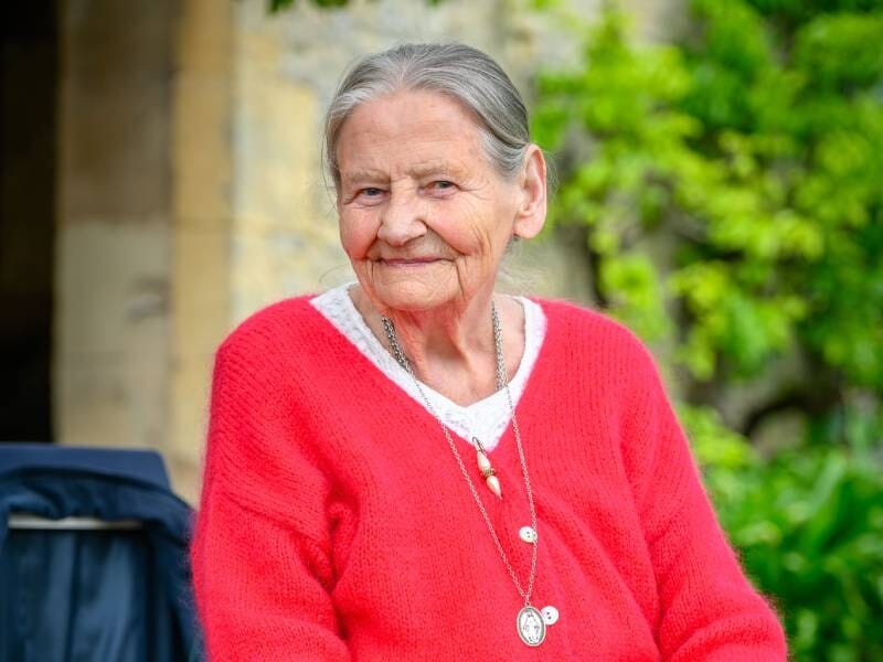 Portrait d'une femme âgée souriante portant un pull rouge, assise dans un jardin avec un mur en pierre en arrière-plan, lors d'un shooting photo près de Rouen.