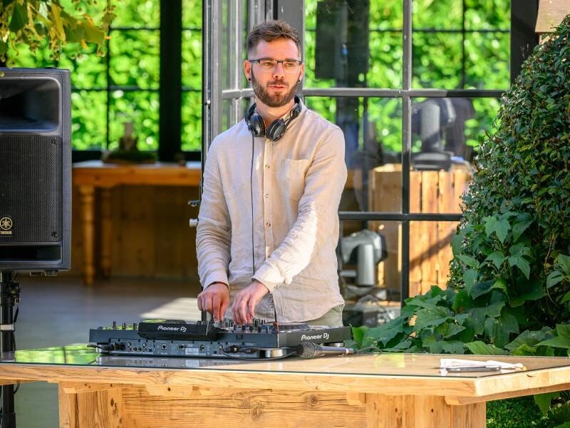 Un homme debout derrière une table en bois, manipulant une console DJ, avec un grand mur vitré et des plantes autour.