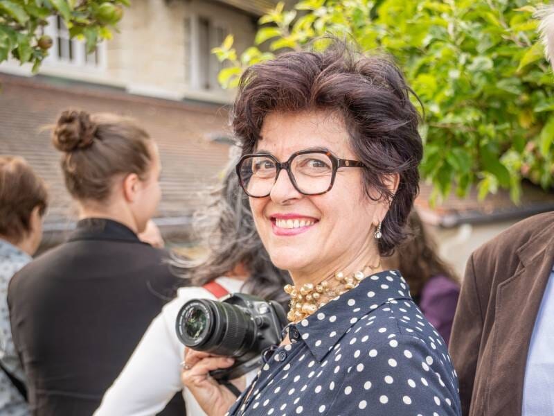 Femme souriante avec lunettes et collier, tenant un appareil photo, entourée de personnes lors d'un événement en extérieur.