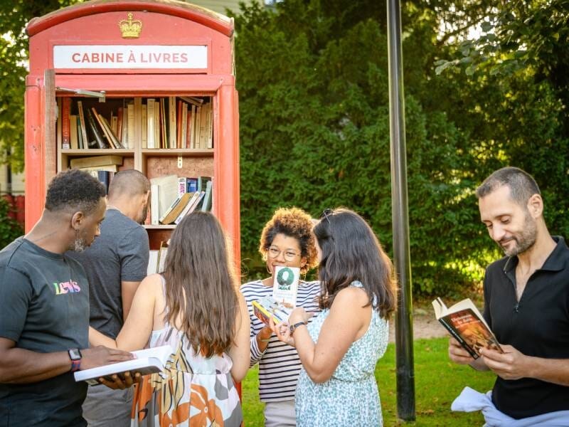 Un groupe de personnes lit des livres devant une cabine à livres rouge en plein air, entourée d'arbres verts.