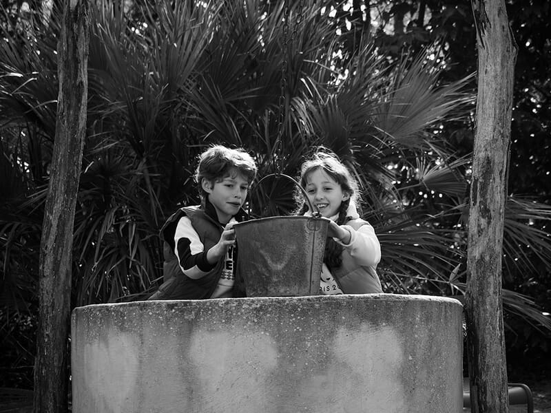 Deux enfants jouent ensemble près d'un puits en pierre, entourés de végétation dense lors d'un shooting photo de famille à Caen.