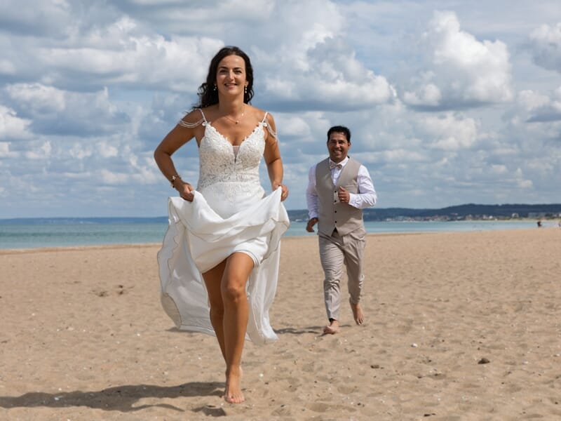 Jeune mariée souriante en robe blanche courant pieds nus sur la plage lors d'un shooting photo à Caen.