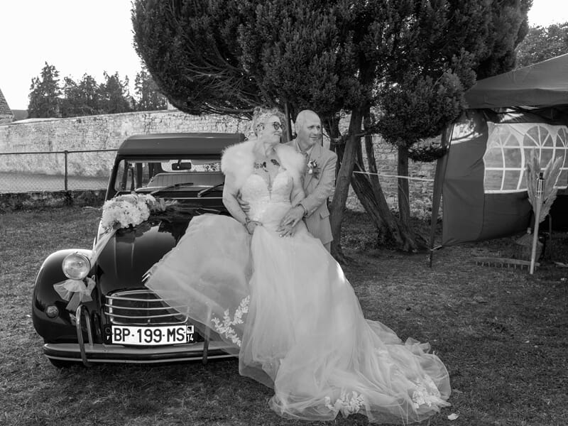 Mariée et marié assis sur le capot d'une voiture ancienne noire décorée de fleurs lors d'un shooting photo à Caen.