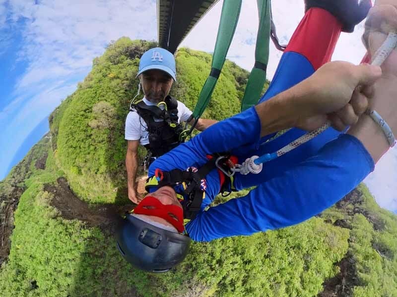 Saut à l'élastique en tandem, vue en plongée sur une vallée verte, avec un instructeur tenant la main du sauteur.