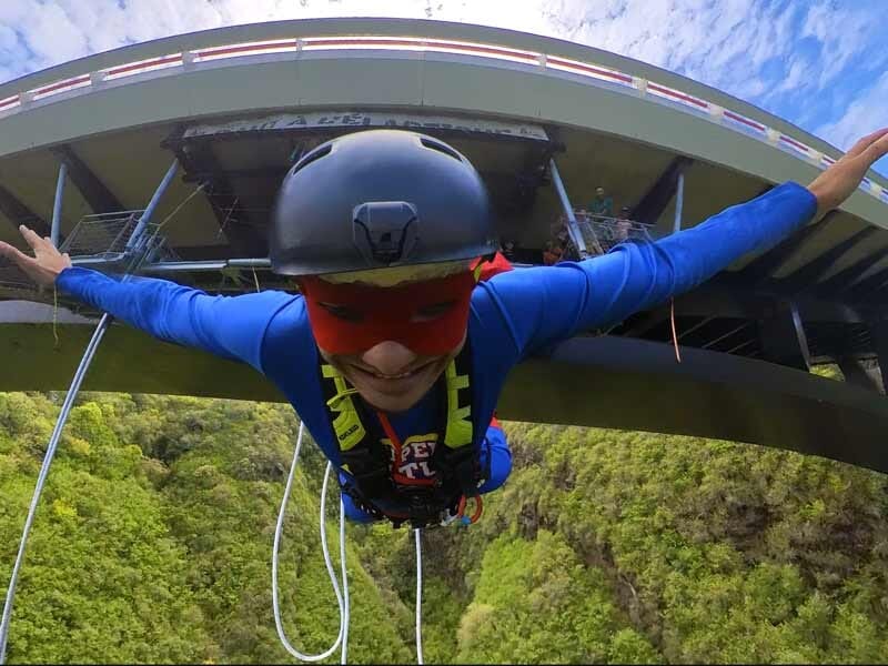 Personne en combinaison bleue effectuant un saut à l'élastique depuis un pont au-dessus d'une vallée verdoyante.