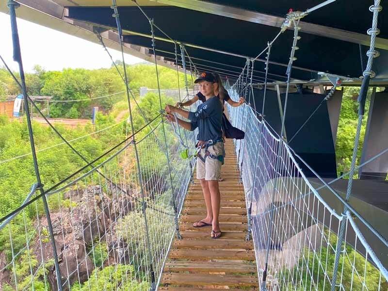Personne debout sur un pont suspendu en bois, tenant les cordes de sécurité, entourée de végétation dense.