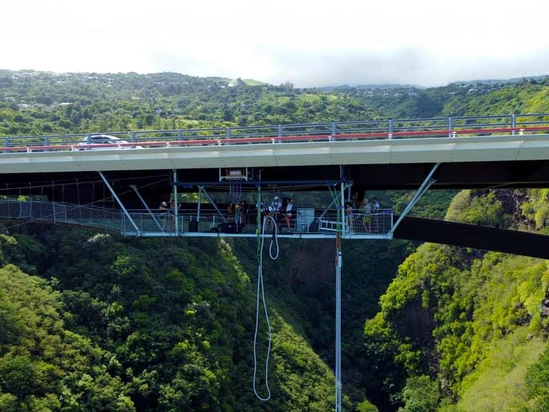 Pont suspendu avec plusieurs personnes et un saut à l'élastique en cours, paysage montagneux et végétation luxuriante.