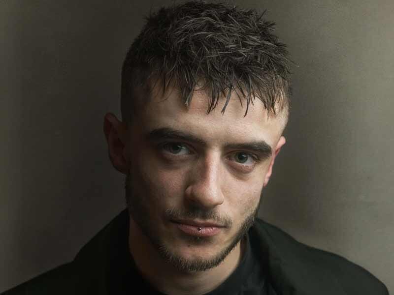 Portrait d'un jeune homme aux cheveux courts et mouillés, regardant l'appareil avec intensité dans un fond neutre.