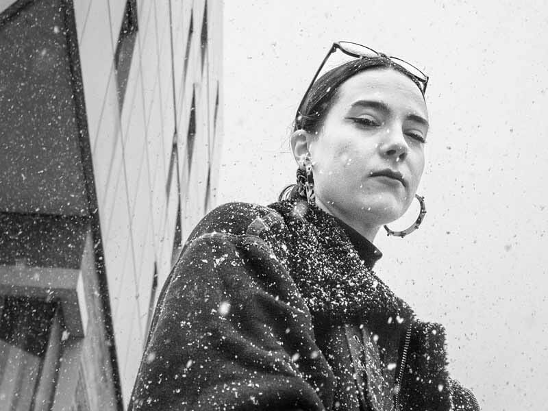 Femme en gros plan sous la neige, portant un manteau sombre et des boucles d'oreilles, avec un bâtiment en arrière-plan.