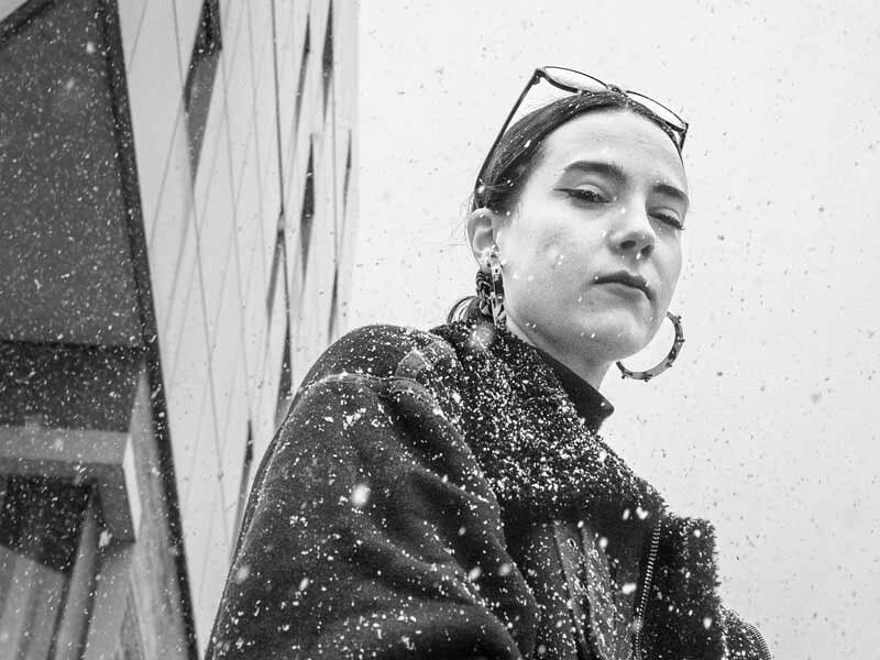 Femme en gros plan sous la neige, portant un manteau sombre et des boucles d'oreilles, avec un bâtiment en arrière-plan.