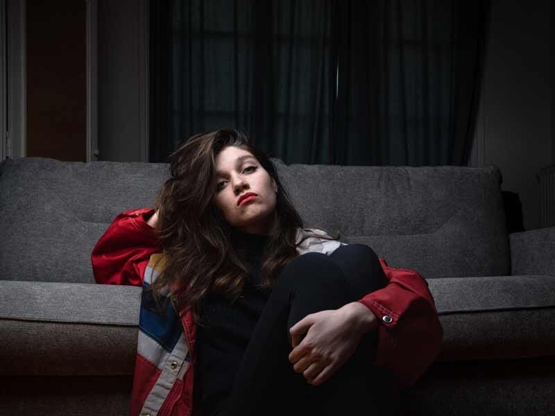Jeune femme assise sur un canapé gris, vêtue d'une veste rouge, posant avec une expression pensive dans un intérieur sombre.