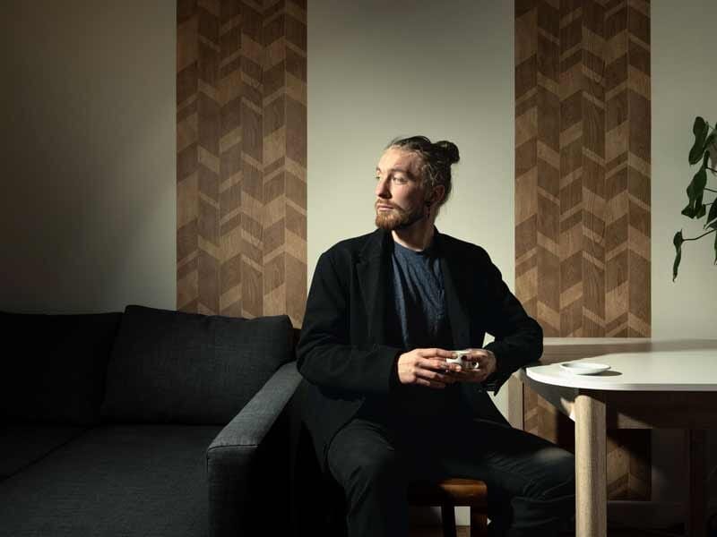 Homme assis sur un canapé dans un salon moderne, tenant une tasse, regardant vers la droite avec une lumière douce.