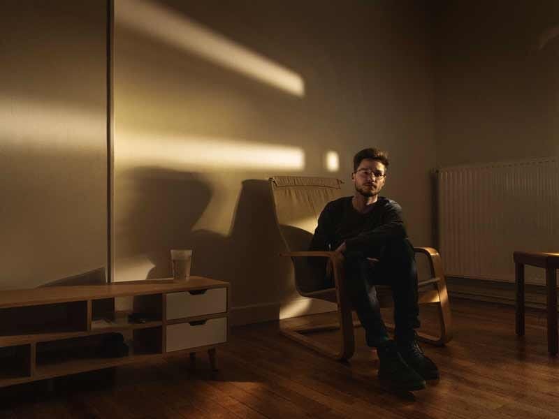 Jeune homme assis sur une chaise dans une pièce éclairée par la lumière du soleil, avec des ombres sur le mur derrière lui.