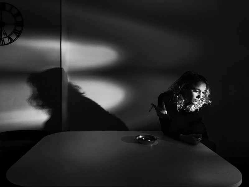 Femme en noir et blanc assise à une table, avec une ombre marquée sur le mur, regardant vers la droite dans une ambiance dramatique.