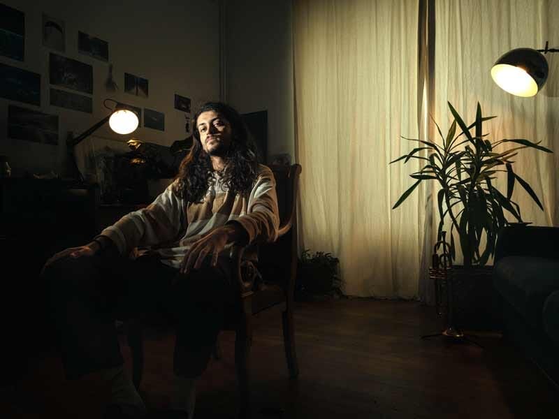 Homme aux cheveux longs assis dans un fauteuil dans une pièce sombre, éclairée par une lampe à côté d'une grande plante verte.